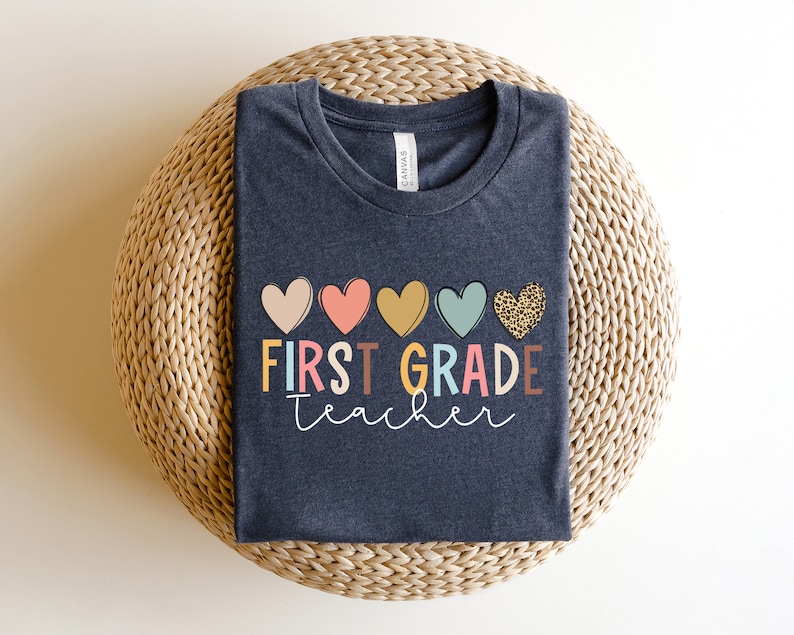 May include: A dark heather grey t-shirt with the text "FIRST GRADE teacher" in colorful letters and a row of five hearts in various colors and patterns above the text.