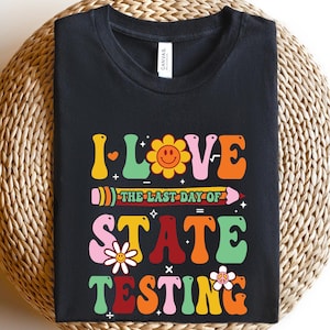 May include: Black t-shirt with a retro-style graphic that says "I Love The Last Day Of State Testing". The graphic features a flower, a pencil, and a heart.