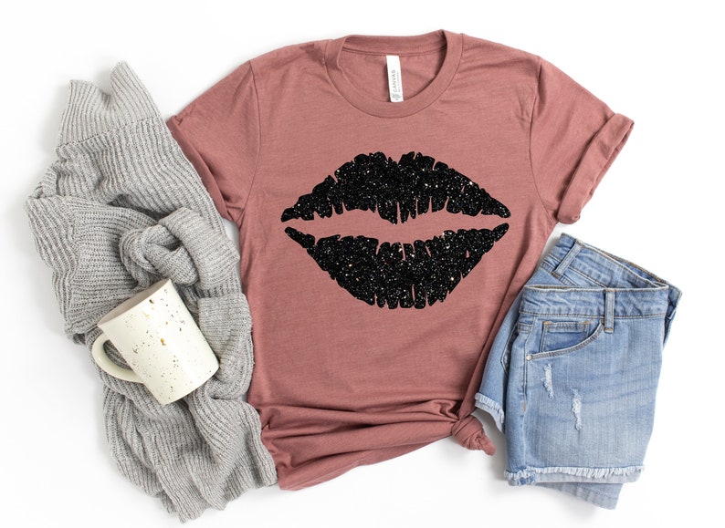 May include: A mauve t-shirt with a black glitter lips design. The t-shirt is shown with a gray sweater and blue denim shorts.