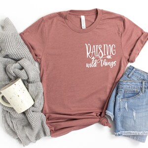 May include: A mauve t-shirt with the text "Raising wild things" in white lettering with an arrow pointing to the right.