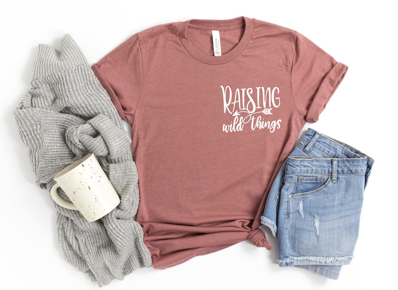May include: A mauve t-shirt with the text "Raising wild things" in white lettering with an arrow pointing to the right.
