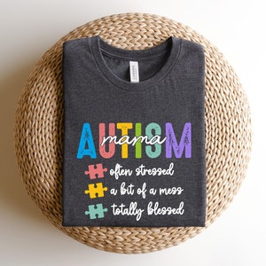 K&ouml;nnte beinhalten: Ein dunkelgraues T-Shirt mit dem Text "AUTISM mama" in Regenbogenfarben. Dem Text folgen drei Puzzle-Teile mit dem Text "oft gestresst", "ein bisschen ein Chaos" und "total gesegnet".