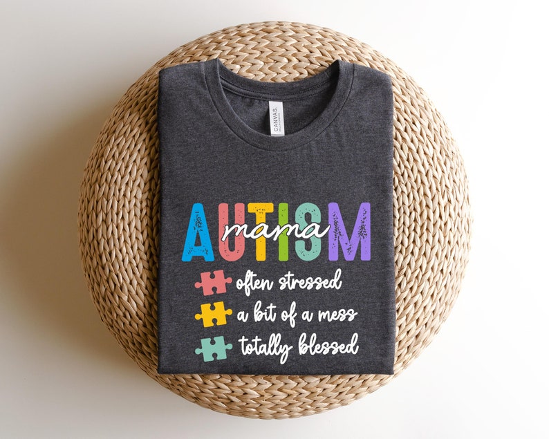 K&ouml;nnte beinhalten: Ein dunkelgraues T-Shirt mit dem Text "AUTISM mama" in Regenbogenfarben. Dem Text folgen drei Puzzle-Teile mit dem Text "oft gestresst", "ein bisschen ein Chaos" und "total gesegnet".