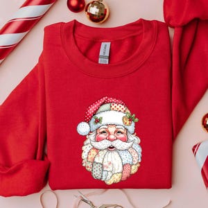 May include: A red sweatshirt featuring a colorful Santa Claus graphic. The Santa Claus design includes a patchwork beard and hat with a polka dot pattern. Christmas-themed apparel.