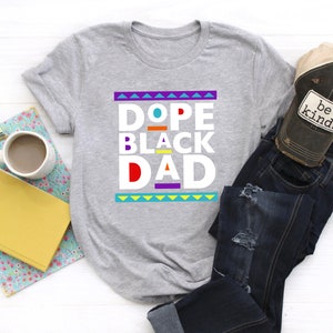 May include: Gray t-shirt with the text "Dope Black Dad" in colorful block letters. The text is surrounded by a colorful geometric pattern.