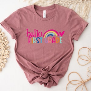 Op de afbeelding: Een mauve t-shirt met een regenboog en de tekst "hello first grade" in kleurrijke letters.