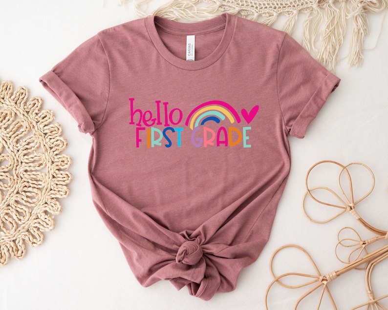 Op de afbeelding: Een mauve t-shirt met een regenboog en de tekst "hello first grade" in kleurrijke letters.