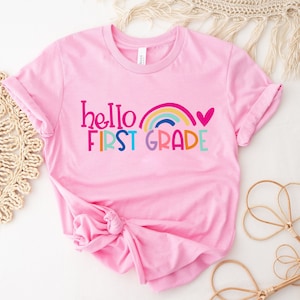 Op de afbeelding: Roze T-shirt met de tekst "hello first grade" en een regenboog met een hart boven de tekst.