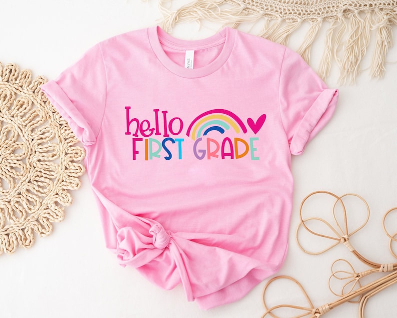 Op de afbeelding: Roze T-shirt met de tekst "hello first grade" en een regenboog met een hart boven de tekst.