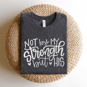 Op de afbeelding: Een donkergrijs T-shirt met de tekst "NOT by MY strength but HIS" in witte letters. Het shirt is gevouwen en wordt getoond op een geweven strooien mat.