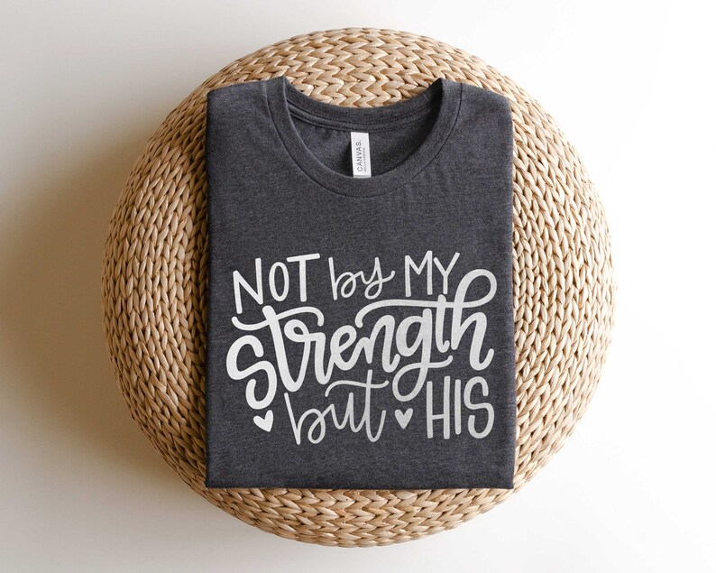 Op de afbeelding: Een donkergrijs T-shirt met de tekst "NOT by MY strength but HIS" in witte letters. Het shirt is gevouwen en wordt getoond op een geweven strooien mat.