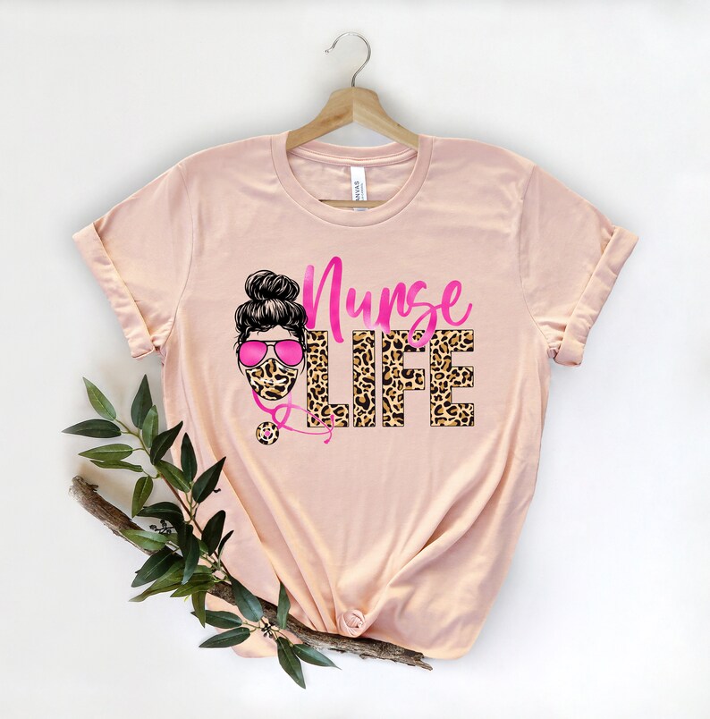 May include: A peach-colored t-shirt with a pink and black leopard print design that says "Nurse Life". The design also includes a cartoon image of a woman wearing a pink face mask and sunglasses.