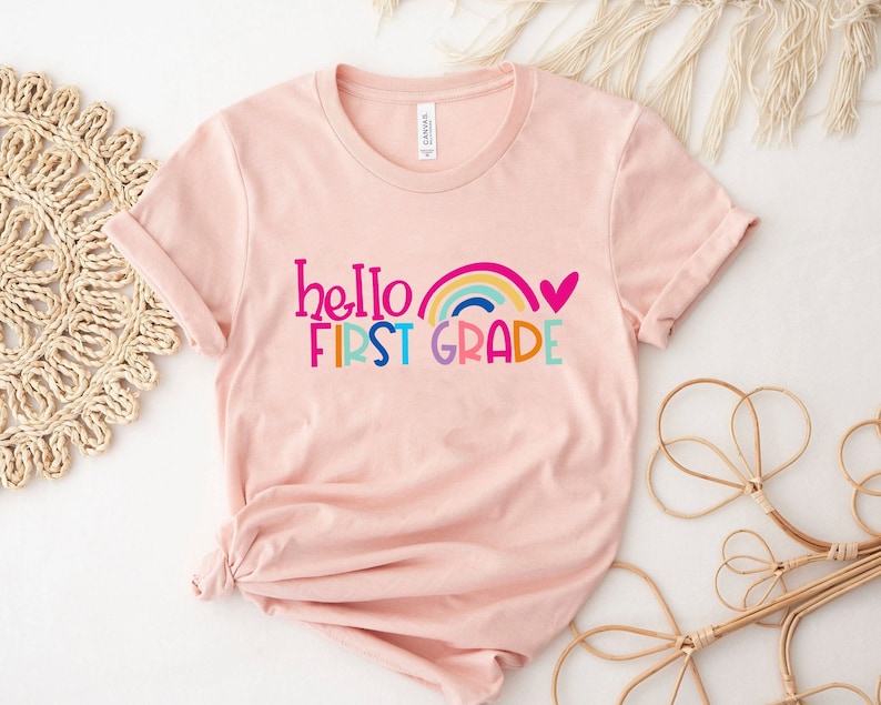Op de afbeelding: Roze T-shirt met de tekst "hello first grade" in kleurrijke letters met een regenboog en een roze hart.