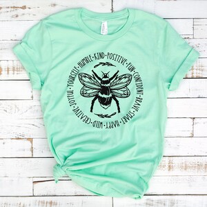 K&ouml;nnte beinhalten: Ein hellgr&uuml;nes T-Shirt mit einer schwarzen Bienengrafik und einem Kreis aus W&ouml;rtern darum herum. Die W&ouml;rter sind: "Wild-Creative-Joyful-Yourself-Humble-Kind-Positive-Fun-Confident-Brave-Smart-Happy."