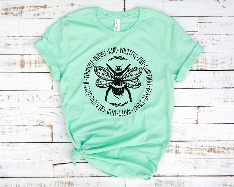 K&ouml;nnte beinhalten: Ein hellgr&uuml;nes T-Shirt mit einer schwarzen Bienengrafik und einem Kreis aus W&ouml;rtern darum herum. Die W&ouml;rter sind: "Wild-Creative-Joyful-Yourself-Humble-Kind-Positive-Fun-Confident-Brave-Smart-Happy."