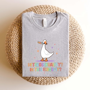 K&ouml;nnte beinhalten: Ein graues T-Shirt mit einer Cartoon-Gans, die einen Strohhut tr&auml;gt und ein St&uuml;ck Brot h&auml;lt. Der Text "Heterosexuality? In this economy?" ist in Regenbogenfarben unter der Gans gedruckt.
