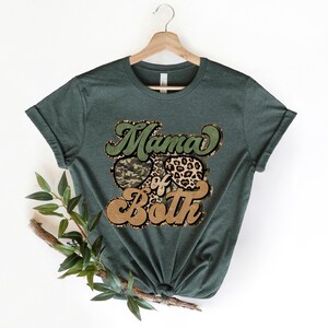 May include: A green t-shirt with the text "Mama of Both" in a retro font. The text is surrounded by a camouflage pattern and a leopard print.