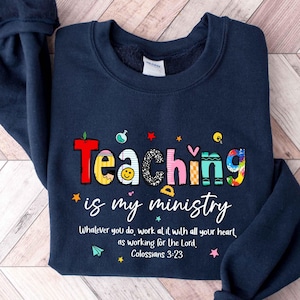 May include: Navy blue sweatshirt with the text "Teaching is my ministry" in colorful letters. The design includes stars, a smiley face, and the Bible verse Colossians 3:23. A comfortable and stylish top for teachers.