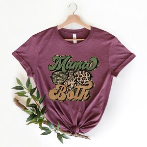 May include: A maroon t-shirt with the text "Mama of Both" in a retro font. The text is surrounded by a camouflage print and a leopard print.