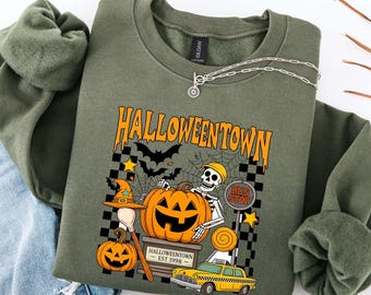May include: Olive green sweatshirt with a Halloween-themed graphic. The design features a jack-o'-lantern, skeleton, bats, and the text "Halloweentown". A yellow taxi cab and a bus stop sign are also included in the design.
