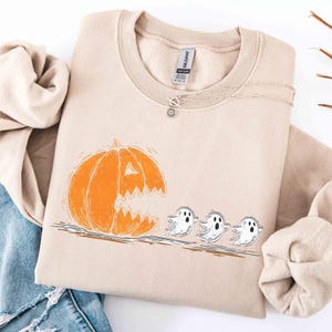 Puede incluir: Una sudadera de cuello redondo color arena con un estampado gráfico de una calabaza naranja grande con boca dentada persiguiendo a tres pequeños fantasmas blancos. La etiqueta de la marca Gildan es visible en el interior del cuello.