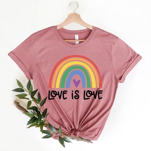 Puede incluir: Una camiseta color malva con un arco&iacute;ris y el texto "LOVE IS LOVE" impreso en la parte delantera.