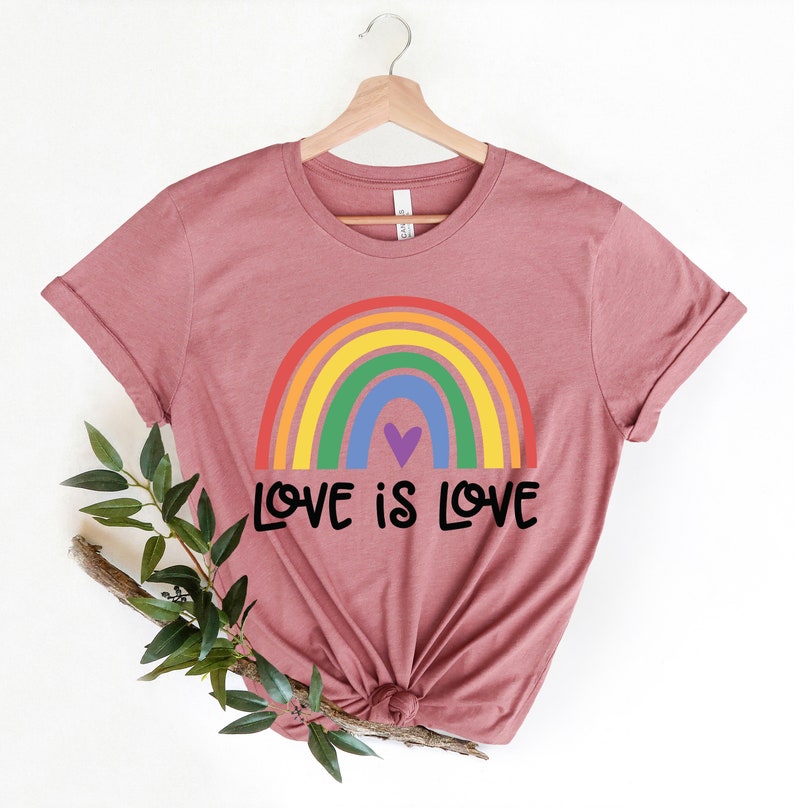 Puede incluir: Una camiseta color malva con un arco&iacute;ris y el texto "LOVE IS LOVE" impreso en la parte delantera.