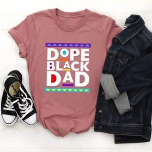 May include: A pink t-shirt with the text "Dope Black Dad" in white letters with colorful accents. The shirt is styled with a denim jacket and black sneakers.