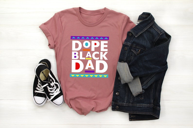 May include: A pink t-shirt with the text "Dope Black Dad" in white letters with colorful accents. The shirt is styled with a denim jacket and black sneakers.