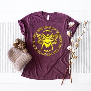K&ouml;nnte beinhalten: Ein maroonfarbenes T-Shirt mit einer gelben Bienengrafik und dem Text "Happy - Wild - Creative - Joyful - Yourself - Humble - Kind - Positive - Fun - Confident - Brave - Smart" um die Biene herum.