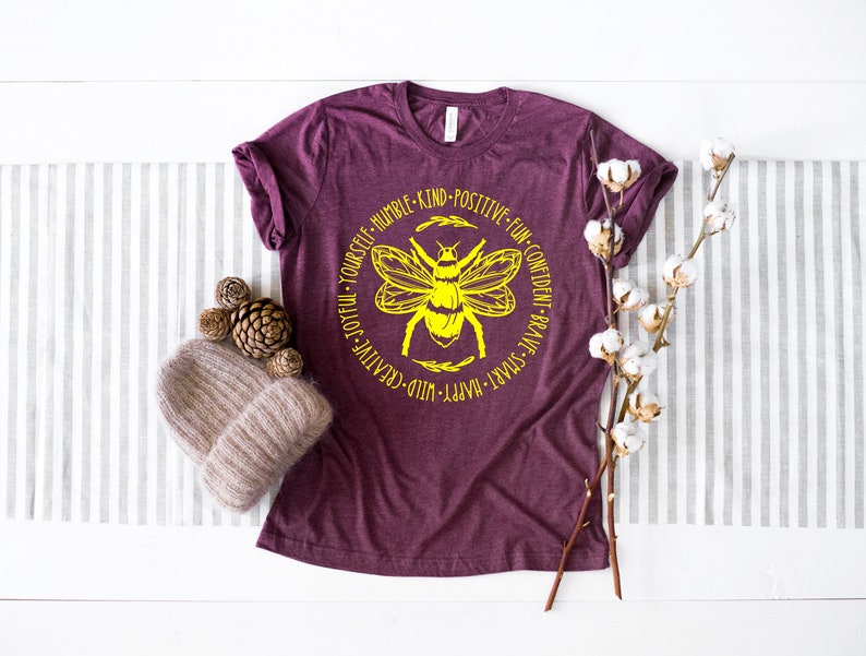 K&ouml;nnte beinhalten: Ein maroonfarbenes T-Shirt mit einer gelben Bienengrafik und dem Text "Happy - Wild - Creative - Joyful - Yourself - Humble - Kind - Positive - Fun - Confident - Brave - Smart" um die Biene herum.
