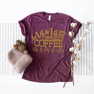 Puede incluir: Una camiseta de color granate con texto en purpurina dorada que dice "mama needs coffee lots & lots of coffee". Se incluye un icono de taza de caf&eacute; en el texto. La camiseta est&aacute; fotografiada sobre una superficie blanca con un camino de mesa a rayas gris y blanco, un gorro de punto marr&oacute;n, pi&ntilde;as y una rama de algod&oacute;n blanco.