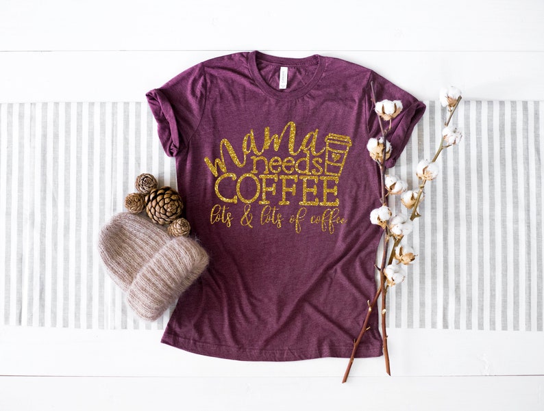 Puede incluir: Una camiseta de color granate con texto en purpurina dorada que dice "mama needs coffee lots & lots of coffee". Se incluye un icono de taza de caf&eacute; en el texto. La camiseta est&aacute; fotografiada sobre una superficie blanca con un camino de mesa a rayas gris y blanco, un gorro de punto marr&oacute;n, pi&ntilde;as y una rama de algod&oacute;n blanco.