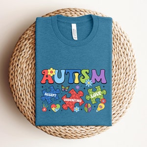 May include: A teal blue t-shirt with the word "AUTISM" in colorful letters. The word is surrounded by puzzle pieces with the words "ACCEPT", "UNDERSTAND", and "LOVE" on them. The puzzle pieces are decorated with flowers and a butterfly.