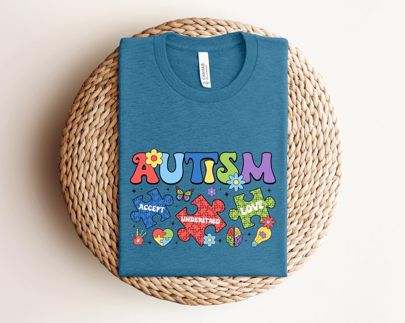 May include: A teal blue t-shirt with the word "AUTISM" in colorful letters. The word is surrounded by puzzle pieces with the words "ACCEPT", "UNDERSTAND", and "LOVE" on them. The puzzle pieces are decorated with flowers and a butterfly.
