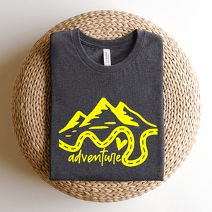 以下が含まれることがあります： 暗いグレーのTシャツに、黄色で山と曲がりくねった道が描かれており、道の終わりにはハートがあります。グラフィックの下には「adventure」という文字が書かれています。