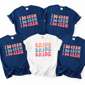 Puede incluir: Un conjunto de camisetas azul marino y blancas. Las camisetas azul marino tienen el texto "I DO CREW" en naranja, azul y rosa. La camiseta blanca tiene "BRIDE" en los mismos colores. Las camisetas son probablemente para una despedida de soltera.