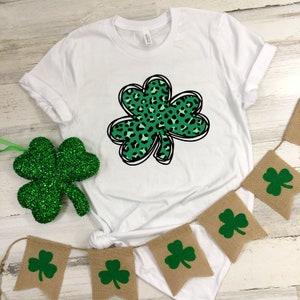 May include: White t-shirt with a green shamrock design featuring a leopard print pattern. The shamrock is outlined in black. The t-shirt is displayed on a white wooden surface with a burlap banner featuring green shamrocks.