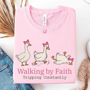 May include: A light pink t-shirt featuring a whimsical design of three cartoon geese with pink bows. The geese are in different poses, with one tripping. The text "Walking by Faith Tripping Constantly" is printed below the geese.