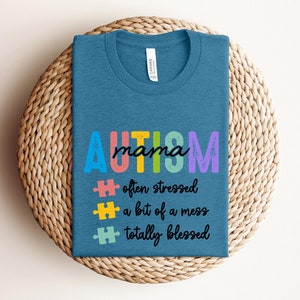 K&ouml;nnte beinhalten: Ein blaues T-Shirt mit dem Text "AUTISM mama" in Regenbogenfarben. Der Text wird von drei Puzzleteilen und dem Text "oft gestresst, ein bisschen ein Chaos, total gesegnet" gefolgt.