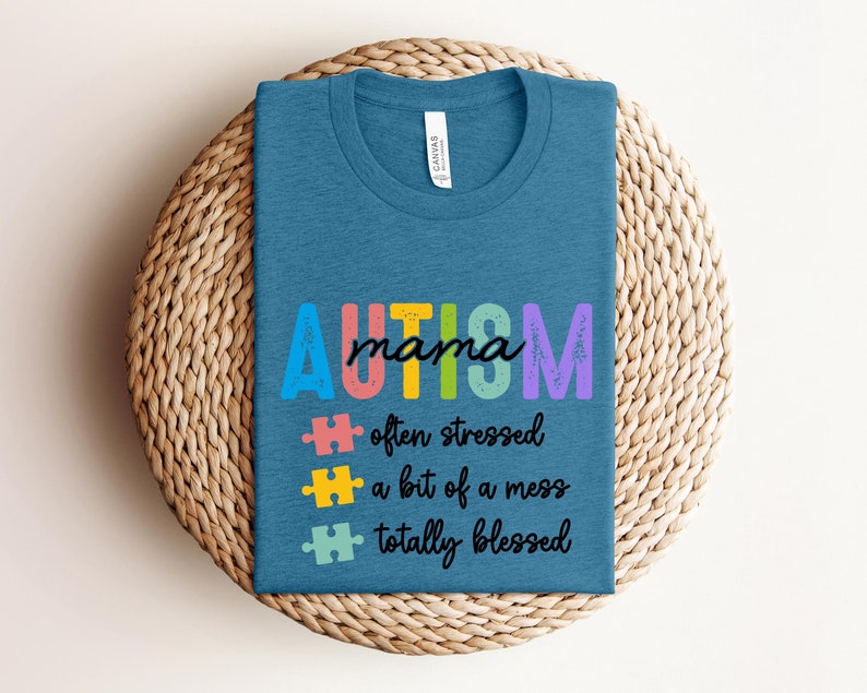 K&ouml;nnte beinhalten: Ein blaues T-Shirt mit dem Text "AUTISM mama" in Regenbogenfarben. Der Text wird von drei Puzzleteilen und dem Text "oft gestresst, ein bisschen ein Chaos, total gesegnet" gefolgt.