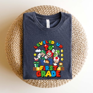 Puede incluir: Una camiseta azul marino con un gr&aacute;fico colorido de Mario, Luigi y otros personajes de la serie de videojuegos Super Mario. El texto "LEVELED UP TO FIRST GRADE" se muestra en una fuente vibrante y multicolor.