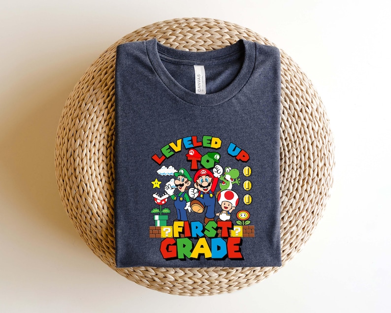 Puede incluir: Una camiseta azul marino con un gr&aacute;fico colorido de Mario, Luigi y otros personajes de la serie de videojuegos Super Mario. El texto "LEVELED UP TO FIRST GRADE" se muestra en una fuente vibrante y multicolor.