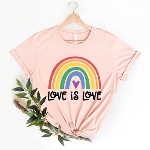 Puede incluir: Una camiseta rosa con un arco&iacute;ris y el texto "LOVE IS LOVE" impreso en ella.