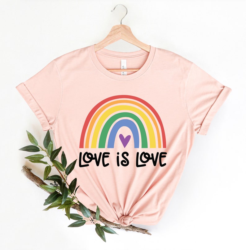 Puede incluir: Una camiseta rosa con un arco&iacute;ris y el texto "LOVE IS LOVE" impreso en ella.
