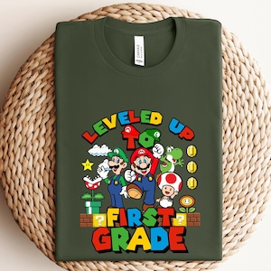 Puede incluir: Camiseta verde oscuro con un gr&aacute;fico colorido de Mario, Luigi y otros personajes del videojuego Super Mario Bros. El texto "LEVELED UP TO FIRST GRADE" se muestra en una fuente vibrante y multicolor.