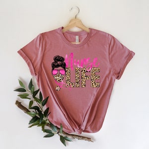 May include: A pink t-shirt with a leopard print design that says "Nurse Life". The design also includes a cartoon image of a woman wearing a leopard print face mask and pink sunglasses.