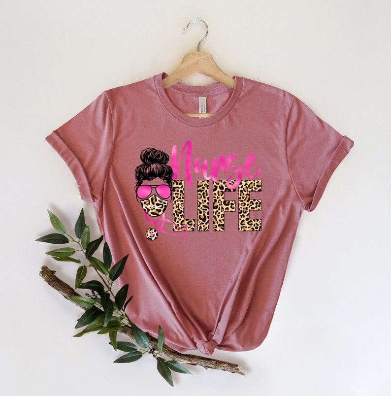 May include: A pink t-shirt with a leopard print design that says "Nurse Life". The design also includes a cartoon image of a woman wearing a leopard print face mask and pink sunglasses.