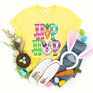 May include: A yellow t-shirt with the words "HOP HOP" in colorful letters and a bunny graphic. The image includes white sneakers, a denim jeans, a carrot, and Easter eggs, suggesting an Easter theme.