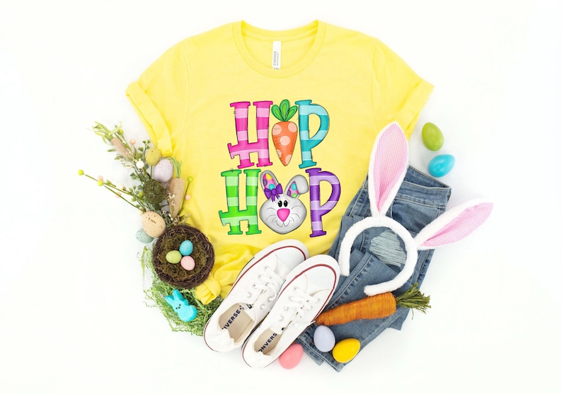 May include: A yellow t-shirt with the words "HOP HOP" in colorful letters and a bunny graphic. The image includes white sneakers, a denim jeans, a carrot, and Easter eggs, suggesting an Easter theme.